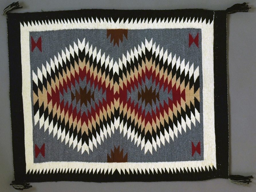 Rectangular woven rug with geometric diamond patterns in black, white, red, brown, beige, and gray, with black fringes on corners.