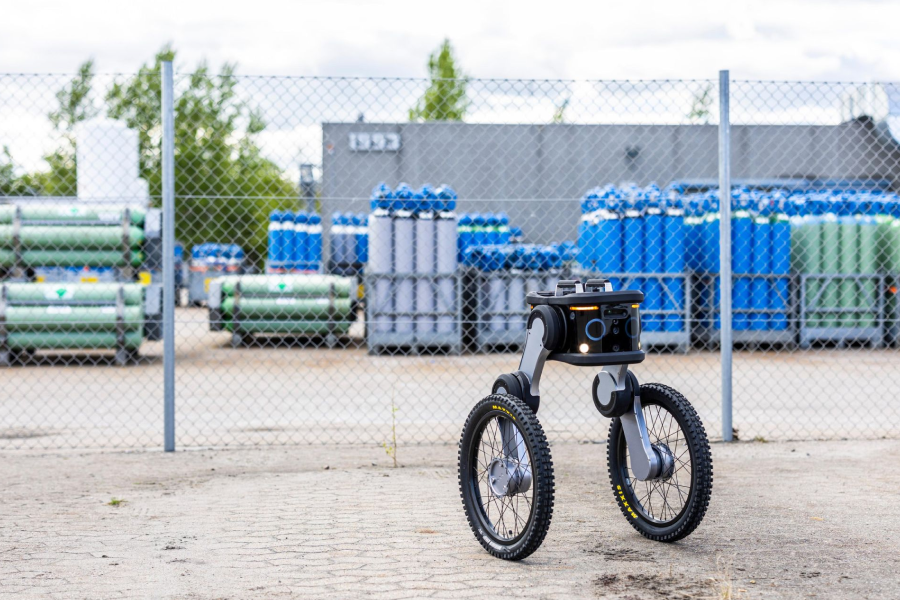 Autonomous security robot patrolling a fenced industrial facility with equipment stored in the background.