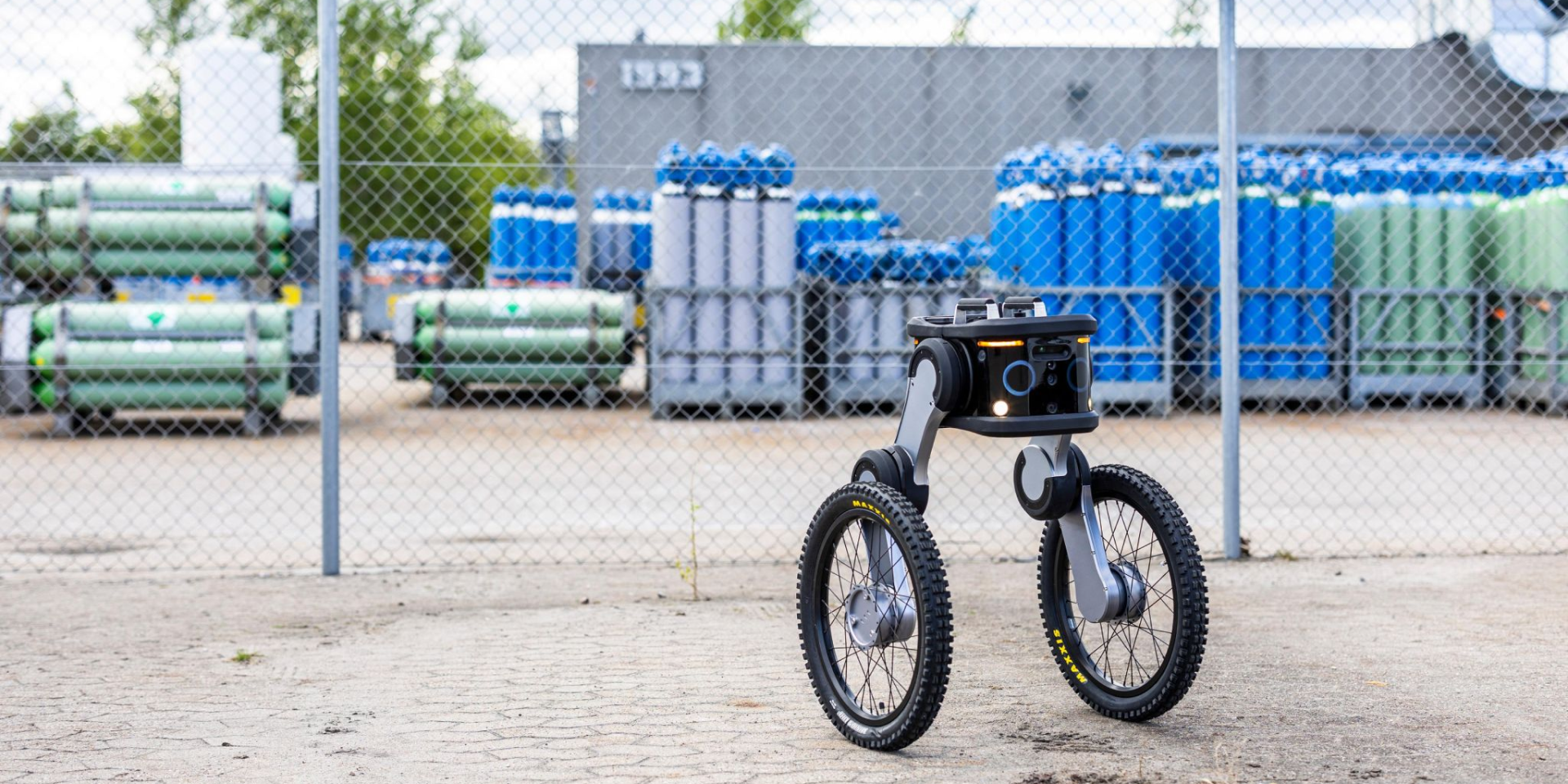 Autonomous security robot patrolling a fenced industrial facility with equipment stored in the background.