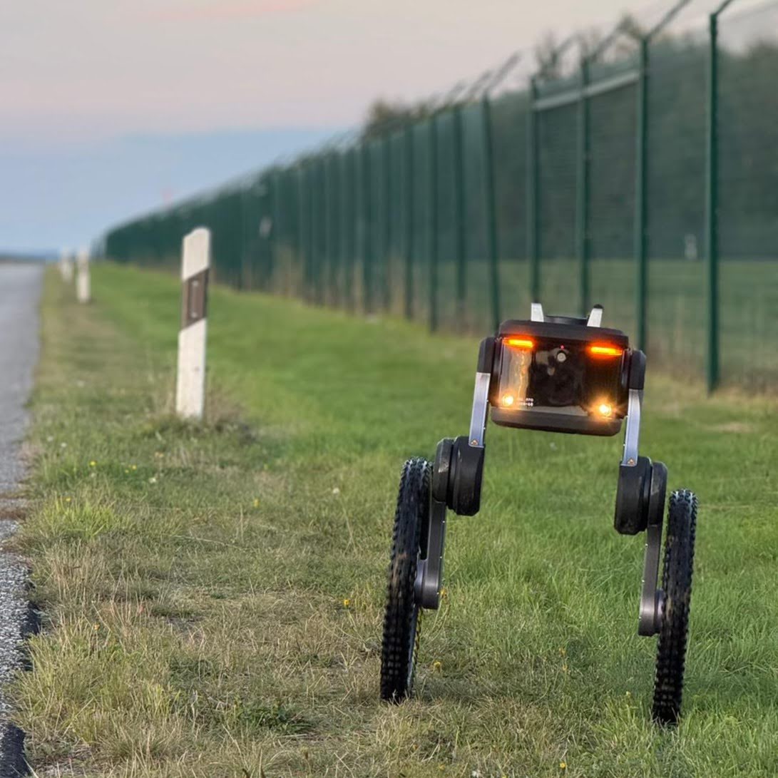 Autonomous security robot patrolling along a fenced perimeter beside an airport roadway.