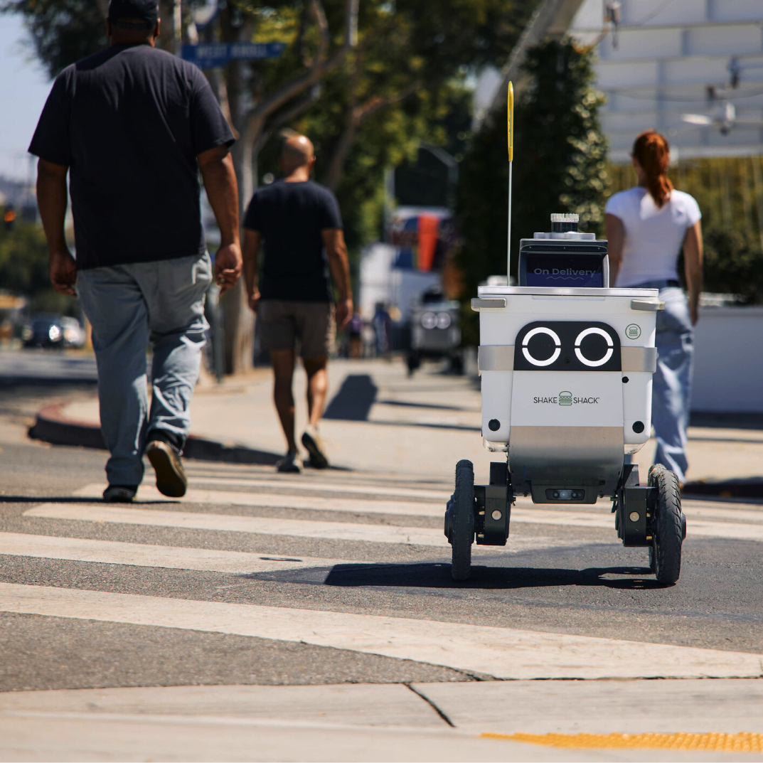 Autonomous food delivery robot traveling on a city street alongside pedestrians.