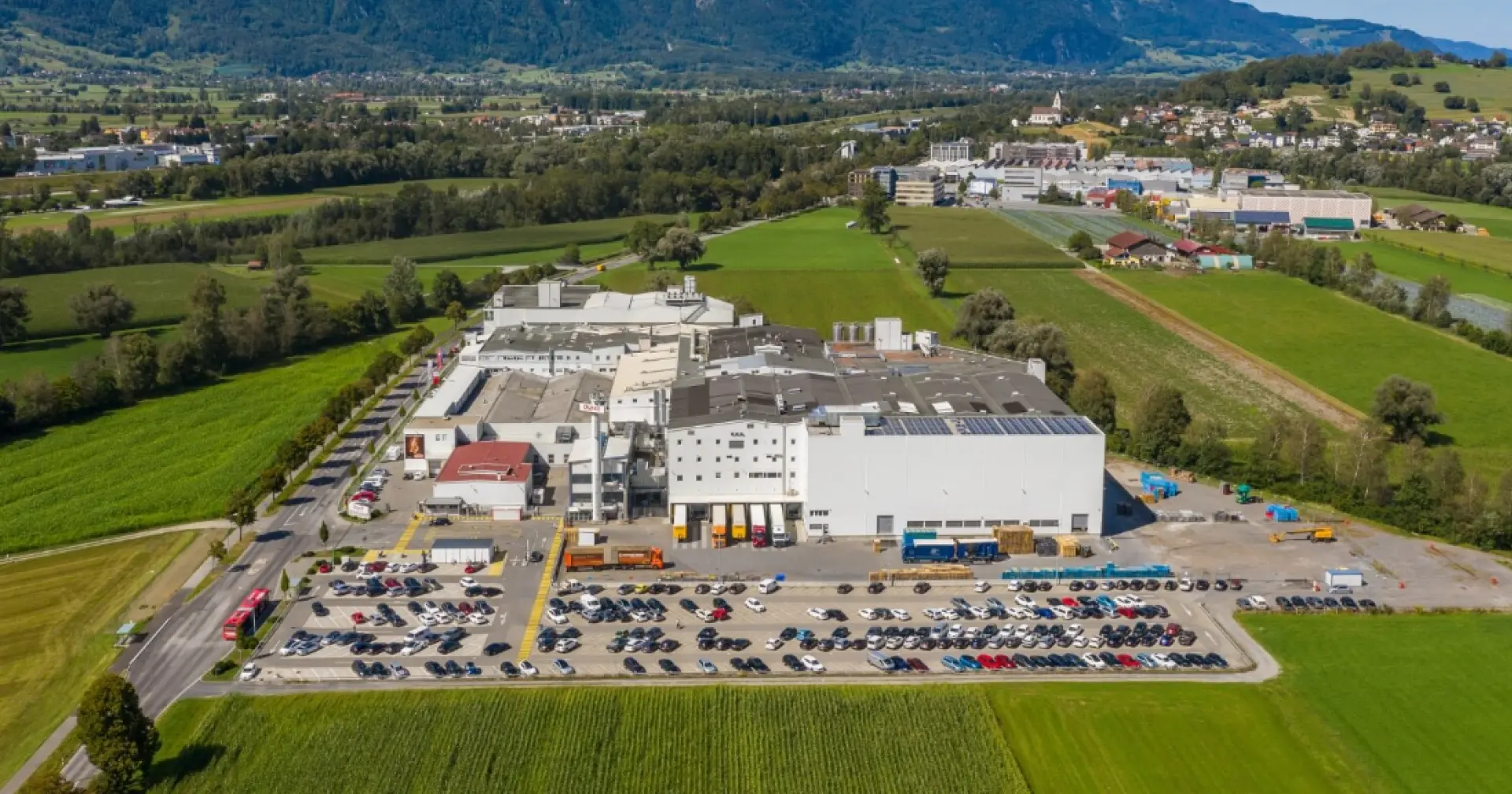 Aerial view of a large industrial manufacturing facility surrounded by fields, parking areas, and nearby buildings.