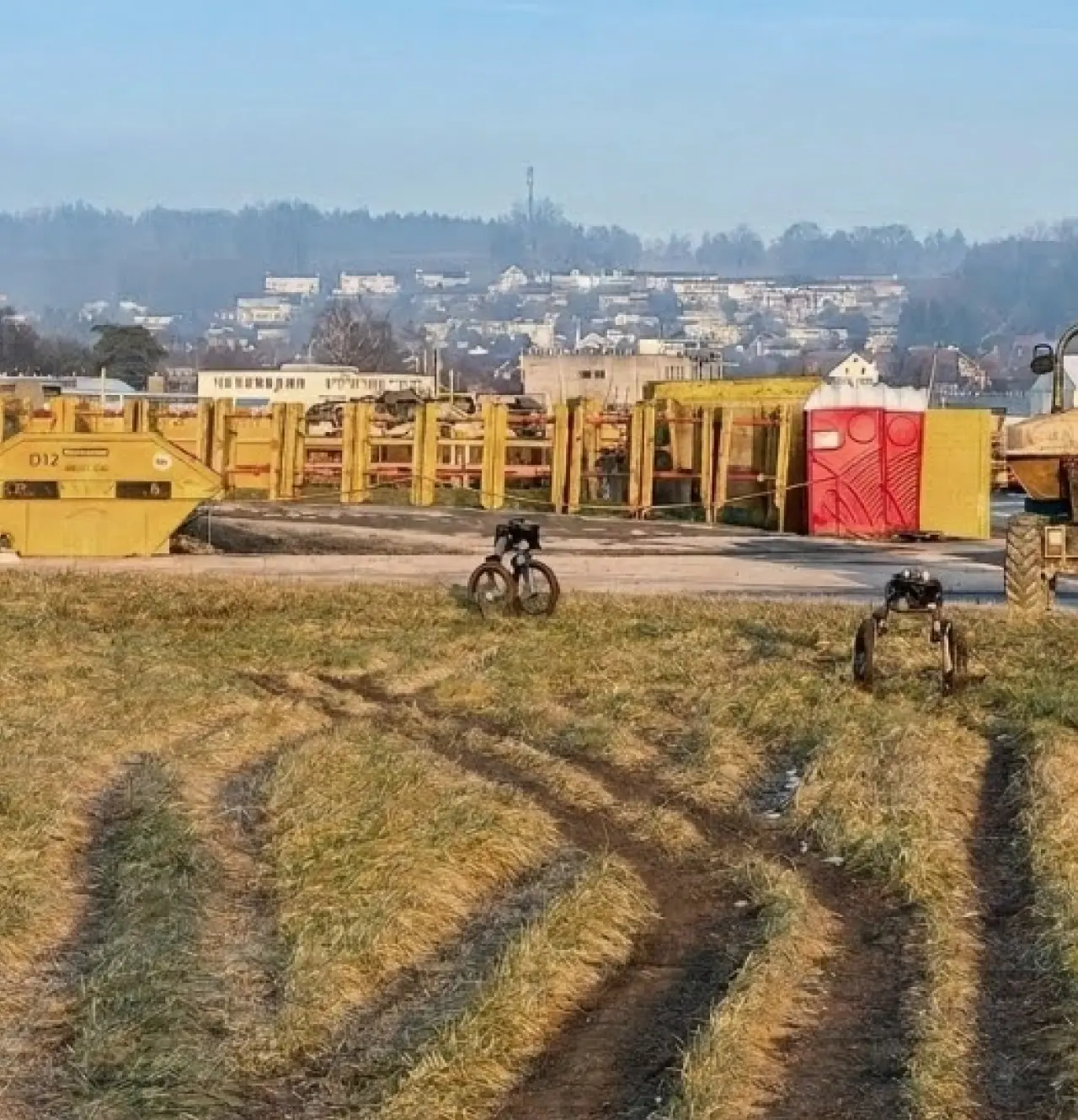 Two autonomous security robots patrolling a grassy industrial site near heavy equipment and perimeter infrastructure.