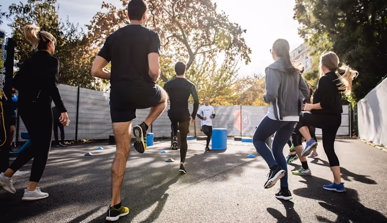 Group of people exercising outdoors by raising knees while facing a trainer near blue cones on a sunny day.