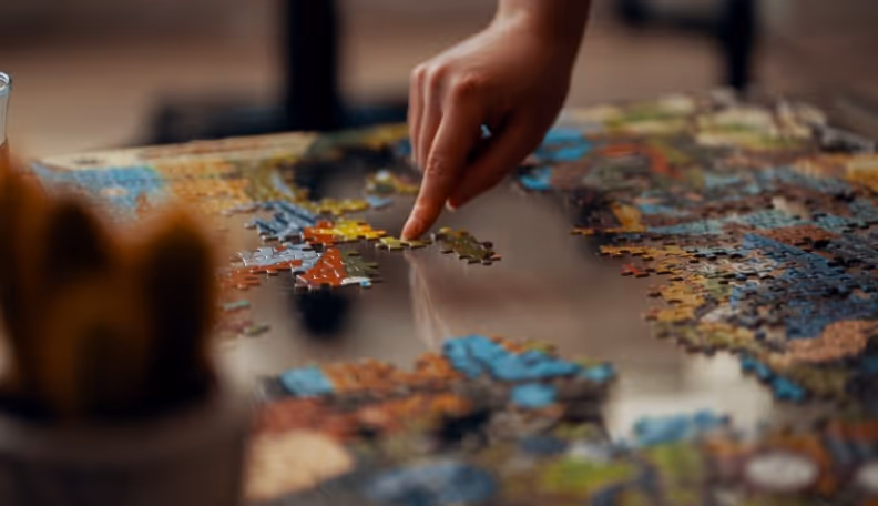 Close-up of a hand placing a puzzle piece on a partially completed colorful jigsaw puzzle on a table.