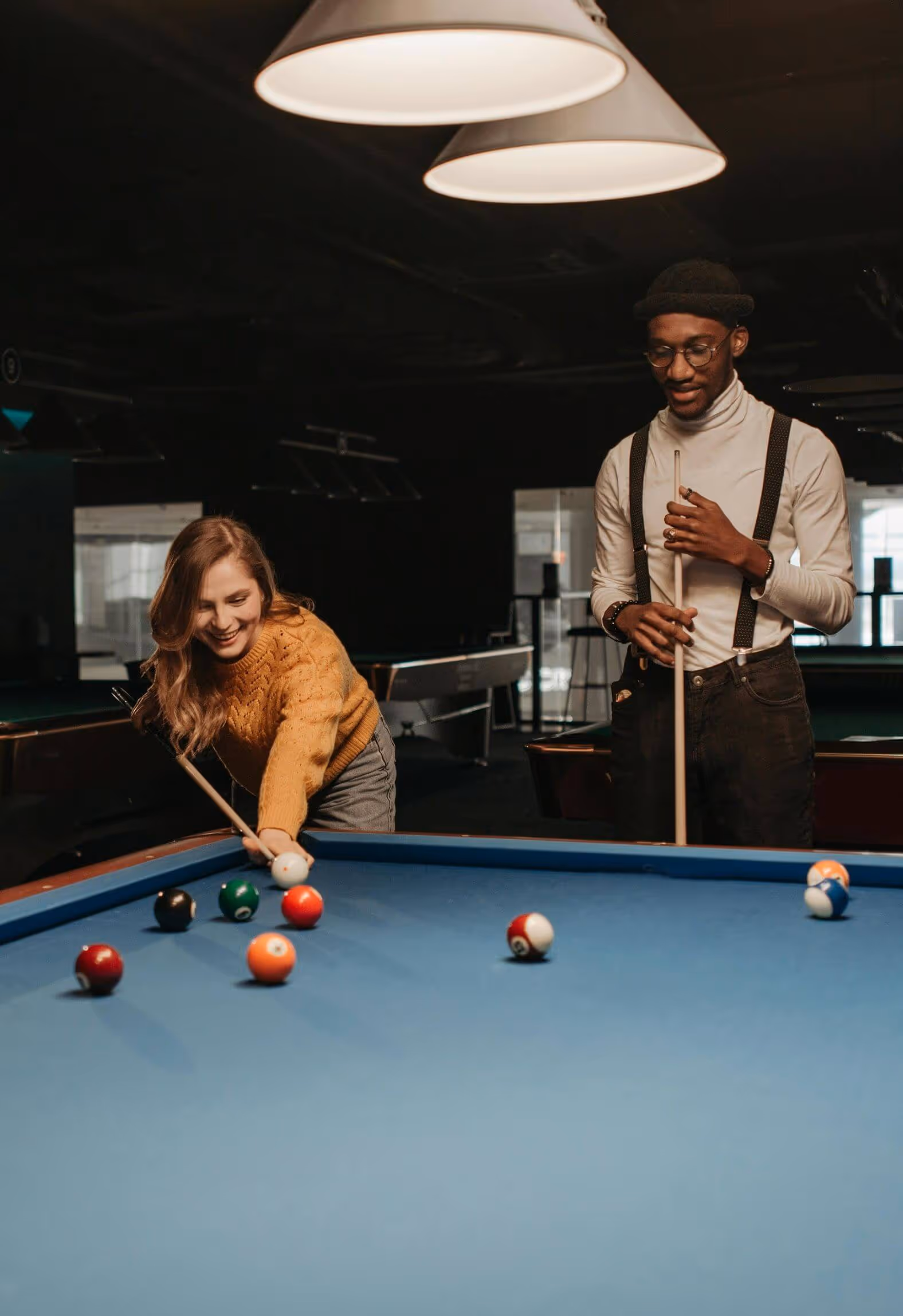 Smiling woman in a mustard sweater aiming a pool cue while a man in glasses, white turtleneck, and suspenders watches nearby.