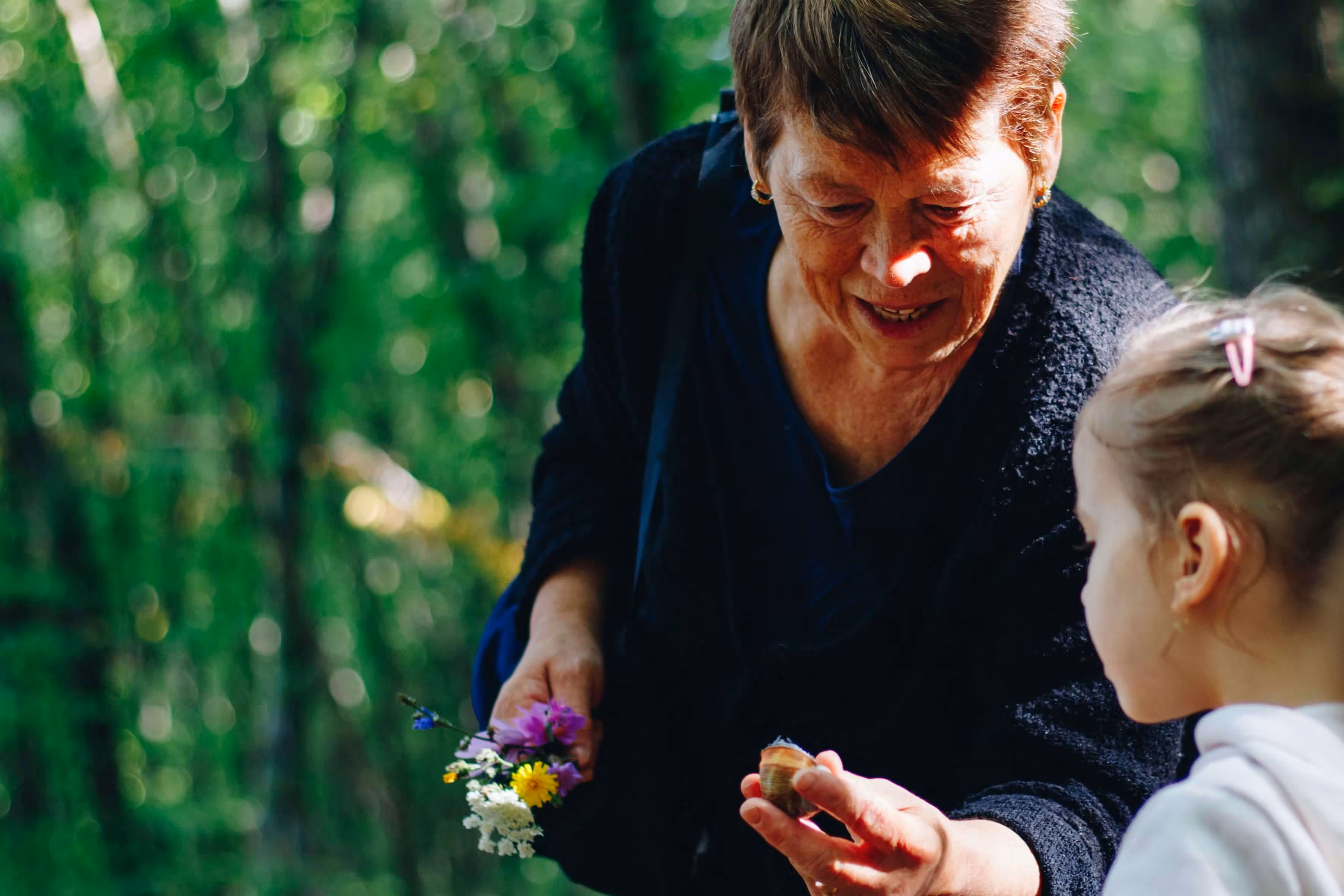 Elderly woman holding flowers and a snail, engaging a young girl in nature learning