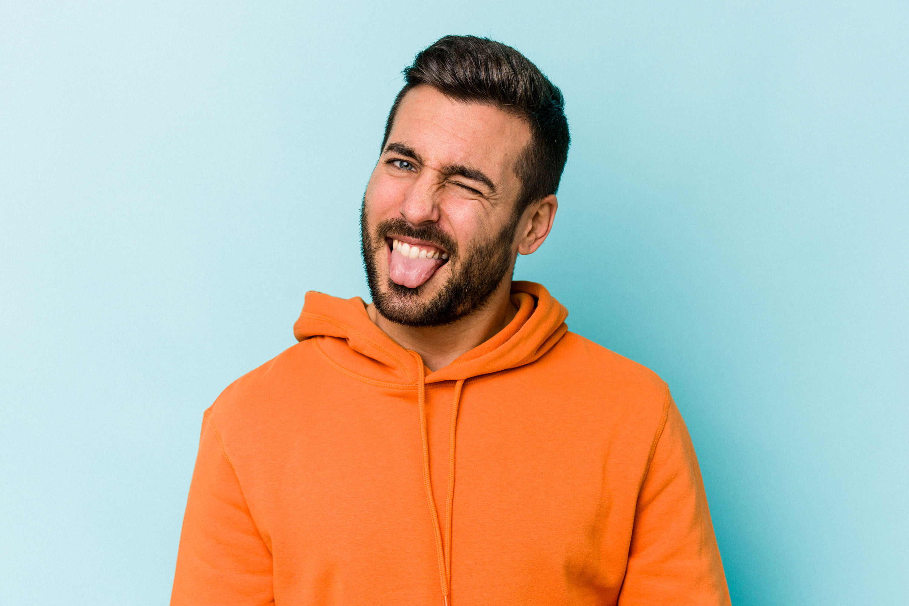 Man in orange hoodie winking with tongue out against a light blue background.