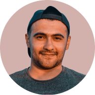 Smiling man with short dark hair wearing a backwards black cap and a gray shirt against a muted pink background.