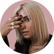 Blonde woman with black nail polish wearing multiple silver rings on her fingers partially covering her face against a muted pink background.