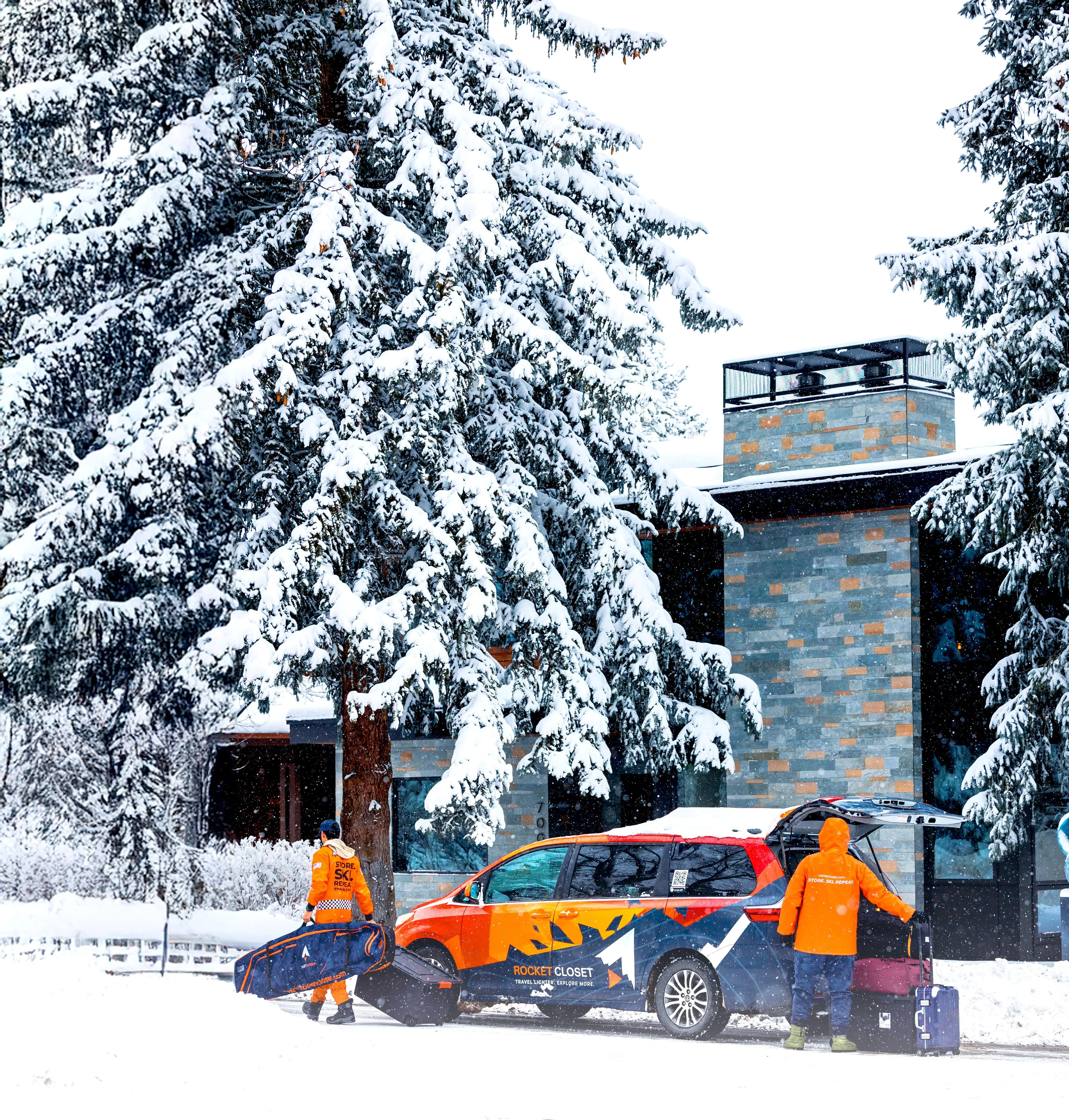 Two people in orange uniforms unloading ski bags from the back of a van in a snowy mountain resort setting.