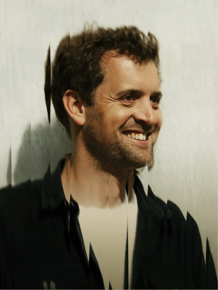 Homme souriant aux cheveux bouclés et barbe légère avec un effet visuel flou vertical.