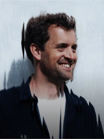 Portrait d'un homme souriant avec cheveux bouclés et barbe légère, fond flou clair.