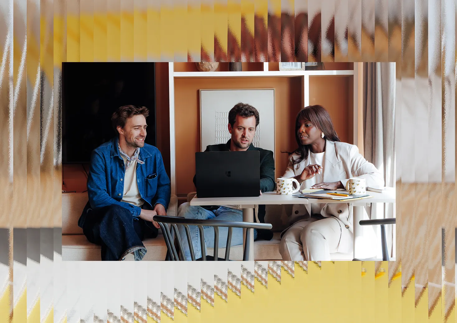 Trois collègues discutent devant un ordinateur portable dans un bureau moderne avec mugs et cahiers.
