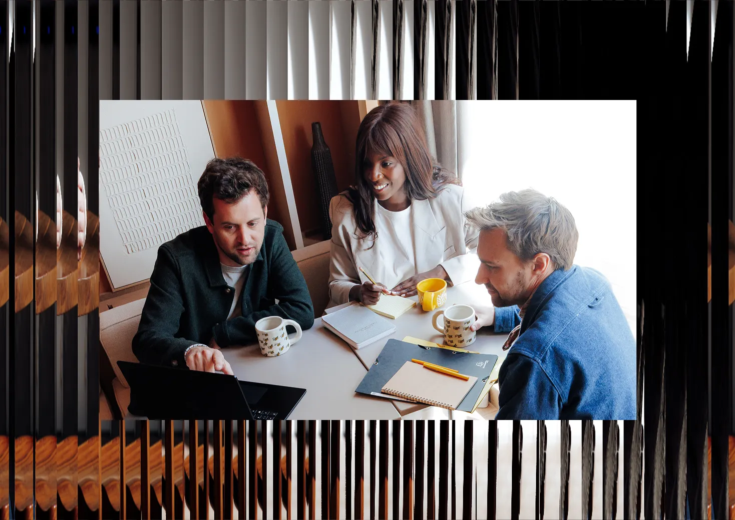 Trois collègues regardant un ordinateur portable avec des tasses, carnets et stylos sur la table.