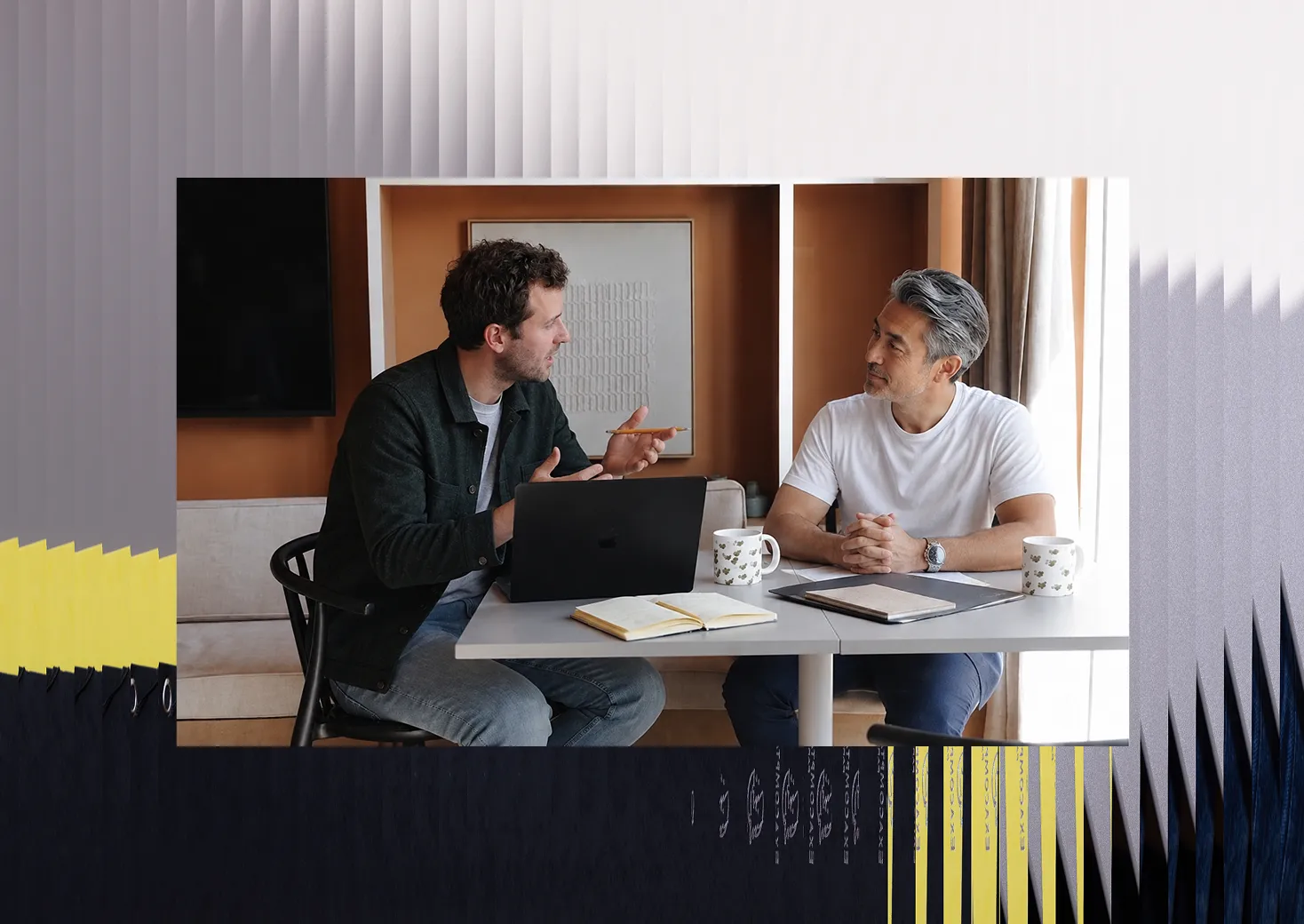 Deux hommes assis à une table en train de discuter, l'un avec un ordinateur portable et un carnet ouvert, l'autre écoutant attentivement, chacun avec une tasse de café.