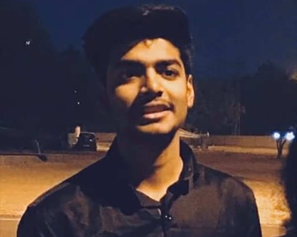 Young man in a black shirt standing outdoors at night with blurred background lights.