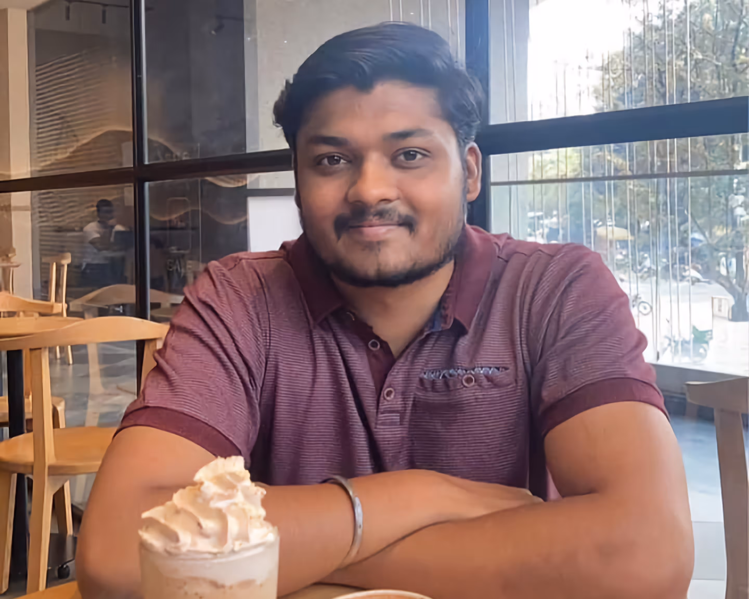 Man sitting at a cafe table with arms crossed, in a maroon polo shirt, smiling at the camera.