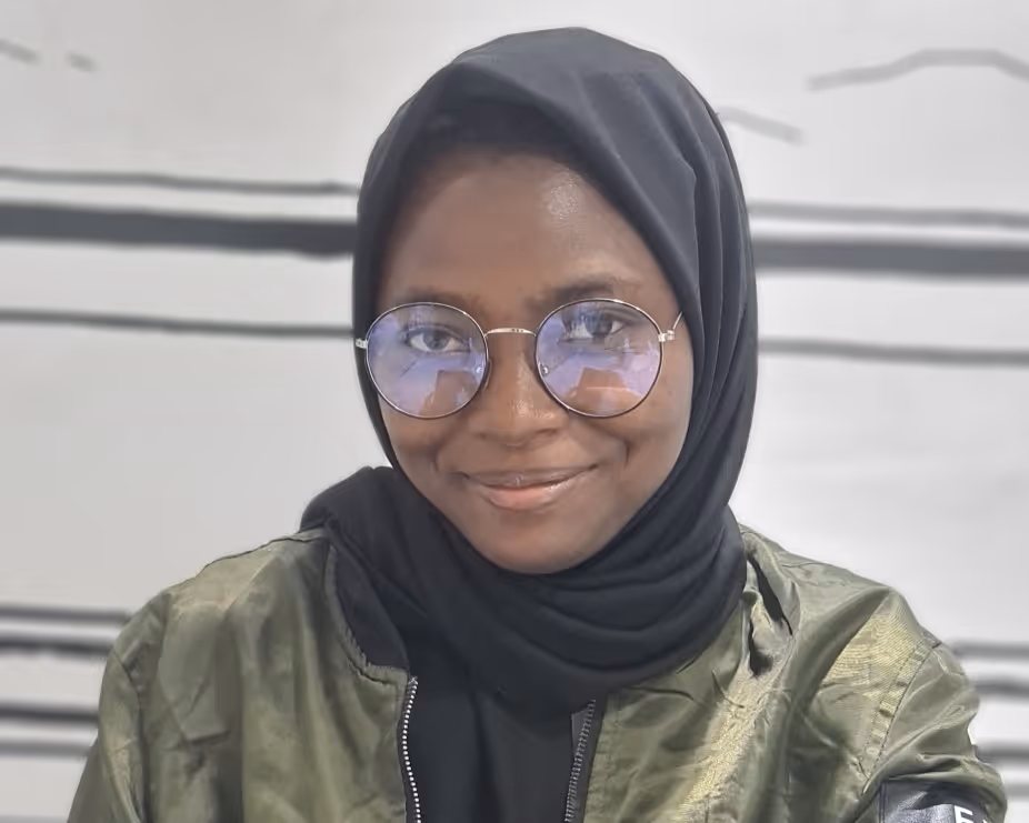 Woman wearing a black hijab and round glasses smiling, with a green jacket and white background.