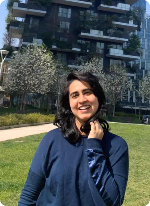 Smiling woman with black hair in blue sweater standing on green lawn near blossoming trees and modern building.
