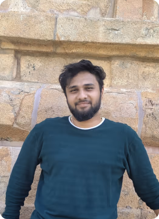 Young man with a beard in a dark green shirt sitting against a stone wall background.