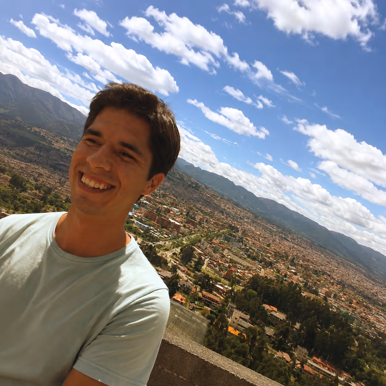 Smiling man in a light blue shirt with a cityscape, trees, mountains, and partly cloudy sky in background.