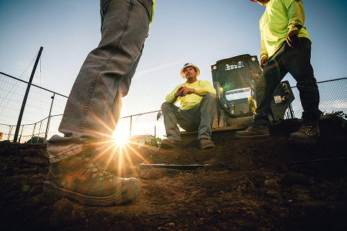 Three construction workers in bright yellow shirts and work boots standing and sitting on dirt at a construction site with the sun low on the horizon behind them.