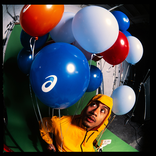 Man in a yellow jacket and hood looking up, surrounded by large colorful balloons in blue, white, red, and orange with a logo, in a studio with lighting equipment and a green backdrop.