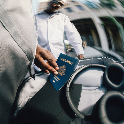 Close-up of a person's hand holding a U.S. passport inside a car, with the person wearing white clothing and sitting behind the wheel.