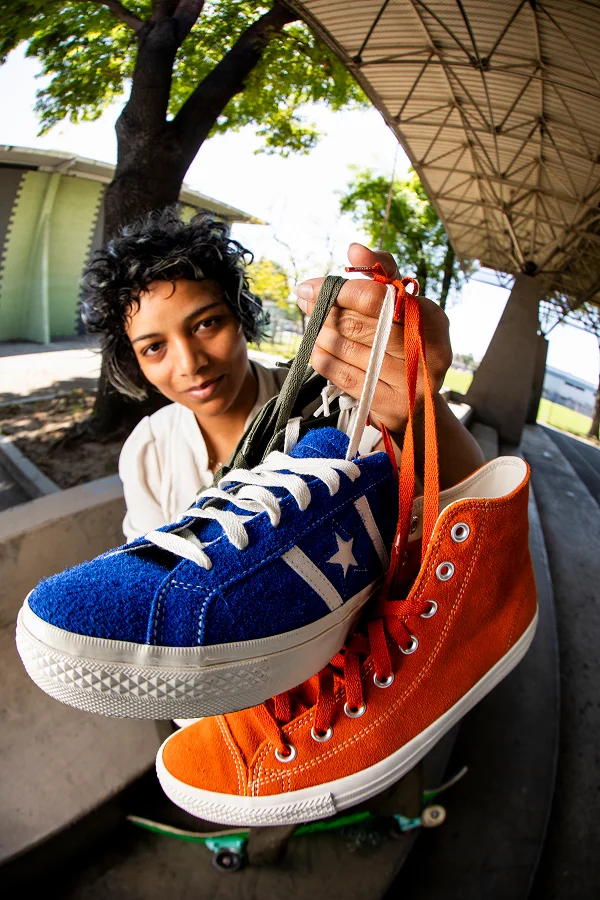 Person holding up two sneakers in front of the camera: a blue low-top sneaker with white laces and a star emblem, and an orange high-top sneaker with orange laces, under a large covered outdoor area with trees in the background.