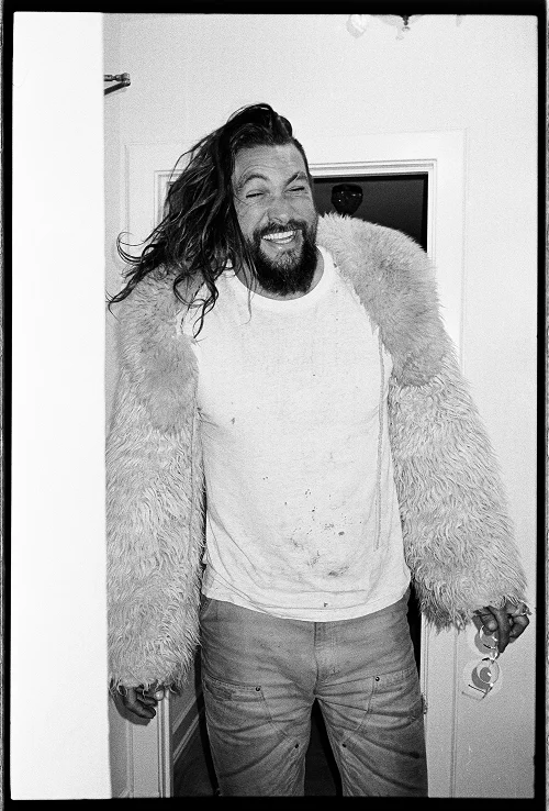 Black and white photo of a man with long hair and beard wearing a fluffy coat over a stained white t-shirt and pants, smiling with eyes partly closed, holding an object in his right hand, standing in a doorway.
