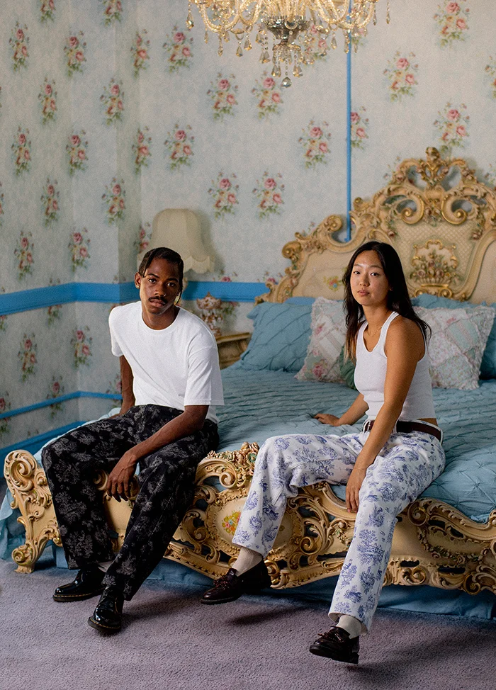 A man and woman sitting on the edge of an ornate vintage bed with a light blue quilt and floral pillows in a room with floral wallpaper and a crystal chandelier. The man wears a white t-shirt and black patterned pants, and the woman wears a white sleeveless top and white pants with blue floral patterns.