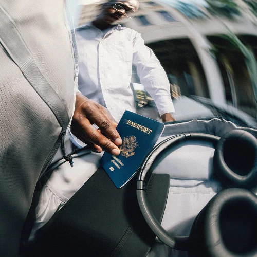 Person holding a blue United States passport while sitting in a modern vehicle with a round steering wheel, wearing a white long-sleeve shirt.