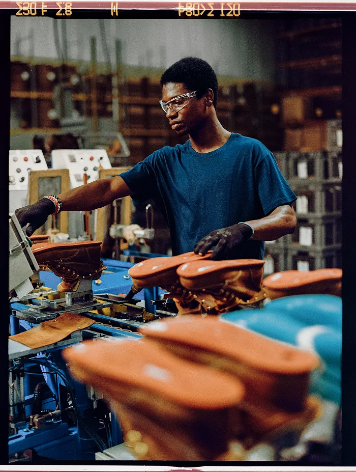 A factory worker wearing safety glasses and gloves inspects brown leather boots on a production line in a manufacturing facility.