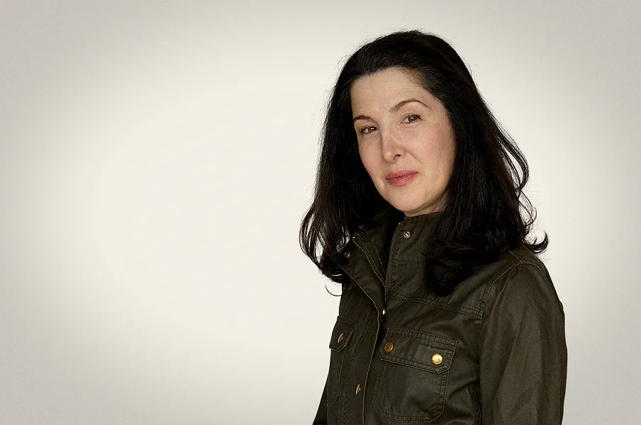 Portrait of a woman with long black hair wearing a dark green jacket, standing against a plain light-colored background, looking at the camera with a subtle smile.