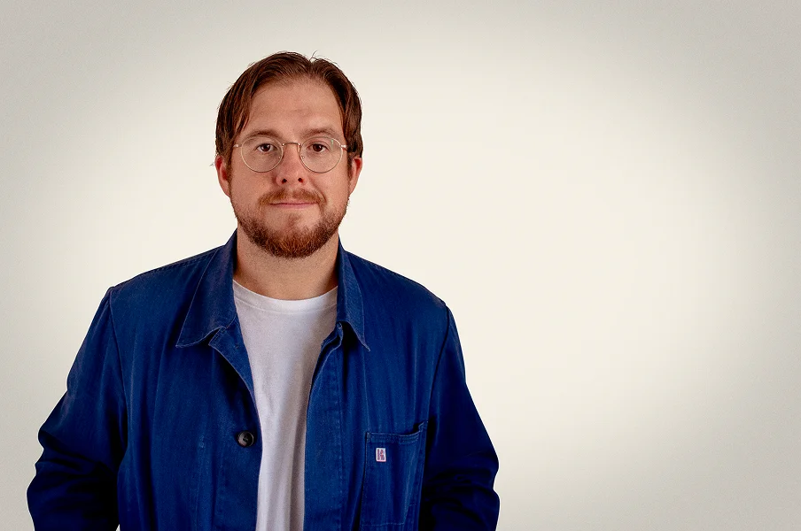 Man with glasses, short brown hair and beard wearing a blue jacket over a white t-shirt, standing against a plain light background.
