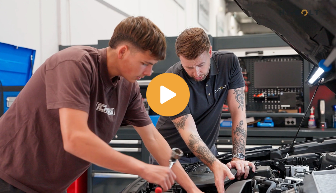 Two men working under the open hood of a car in a workshop, one holding a tool and the other pointing at the engine.