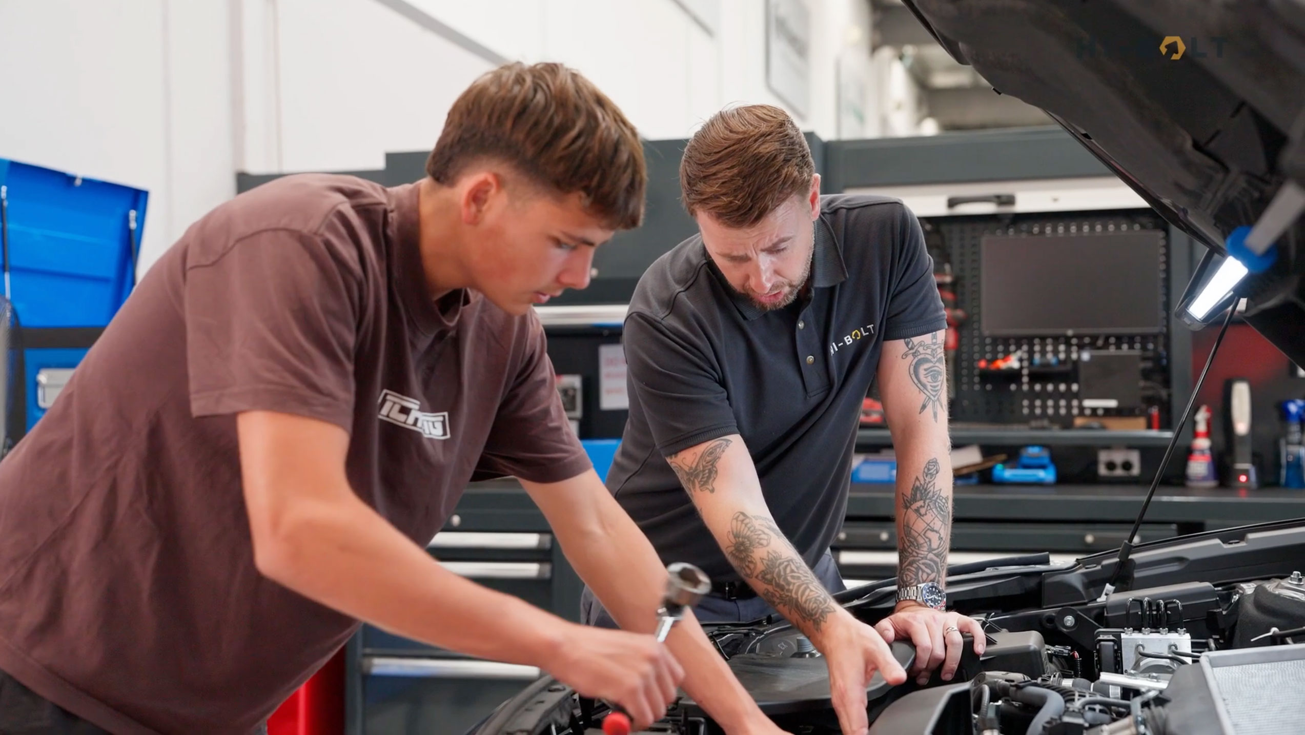 Two men working under the open hood of a car in a workshop, one holding a tool and the other pointing at the engine.