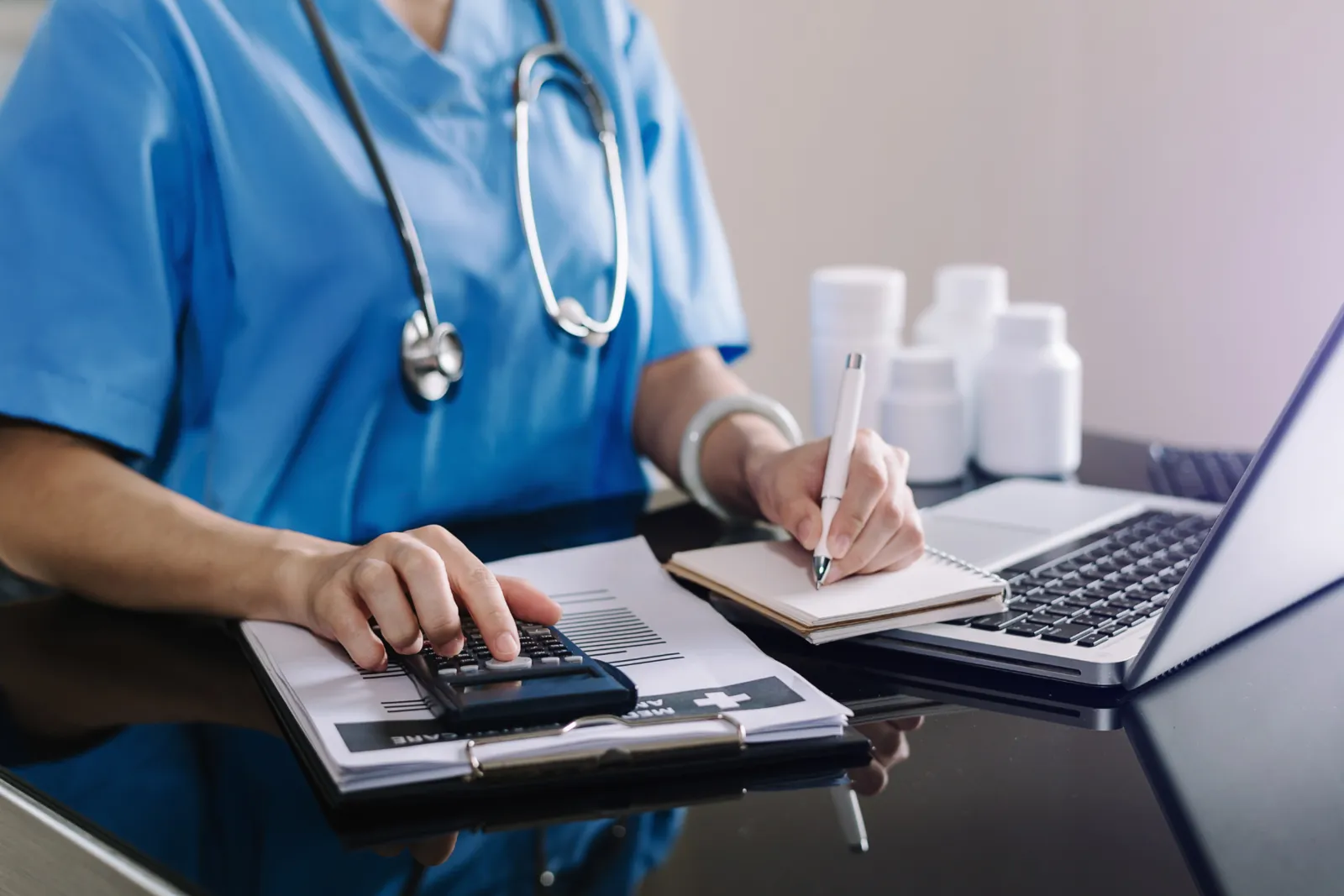 Healthcare worker in blue scrubs working through bills with a calculator and laptop