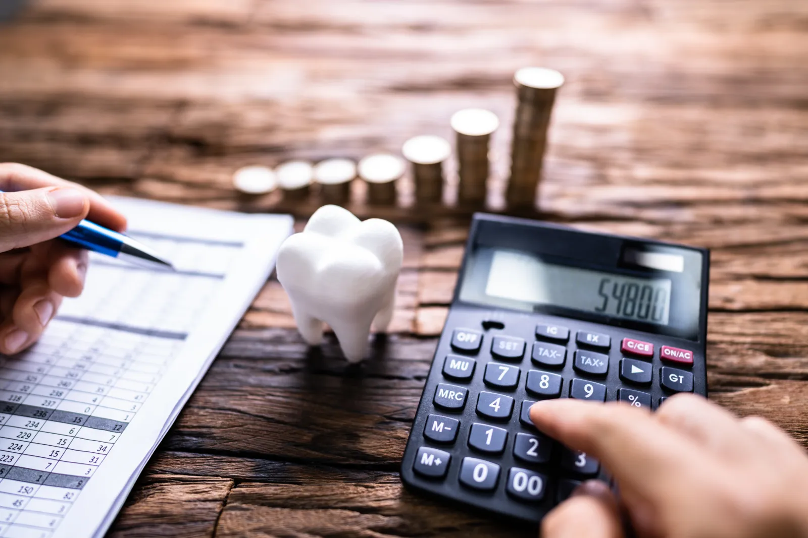 Hand using a calculator beside a tooth-shaped piggy bank and stacks of coins