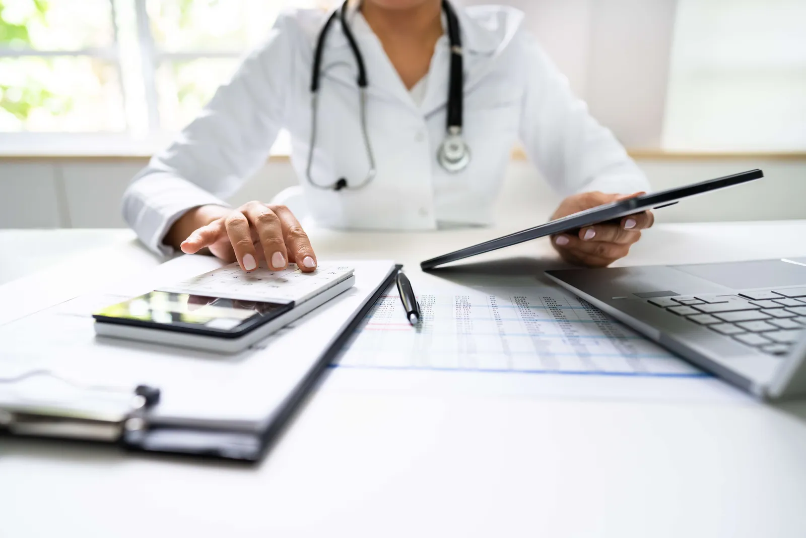 Doctor with a stethoscope working on a tablet and laptop at a desk