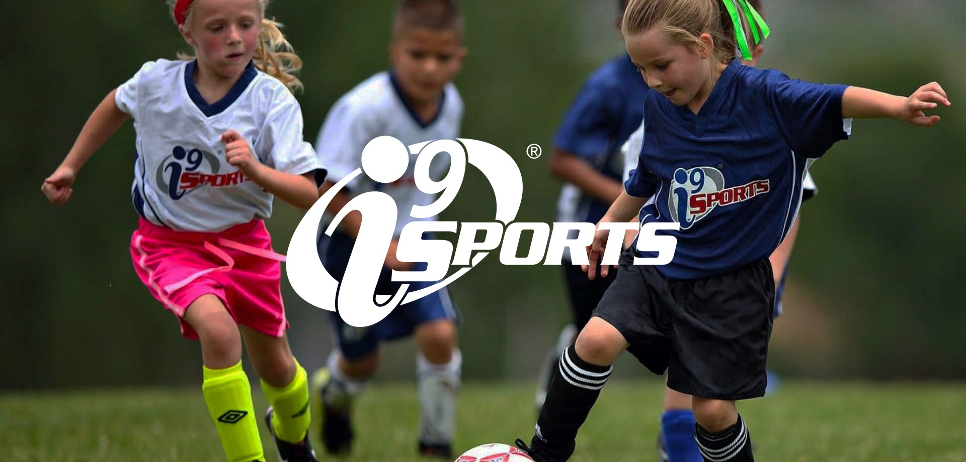 Young children playing soccer on a grass field, wearing uniforms with the i9 Sports logo, with two girls in focus chasing the ball.