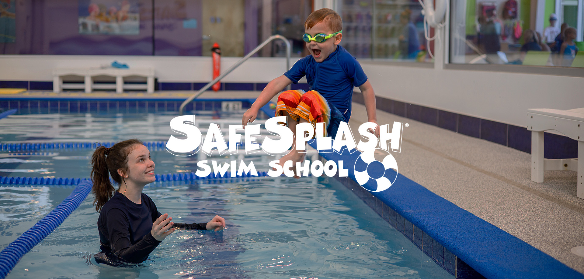 A young boy wearing green goggles and colorful swim trunks jumps excitedly into an indoor pool where a smiling female instructor in a black swim shirt awaits him, with 'SafeSplash Swim School' logo overlaid.
