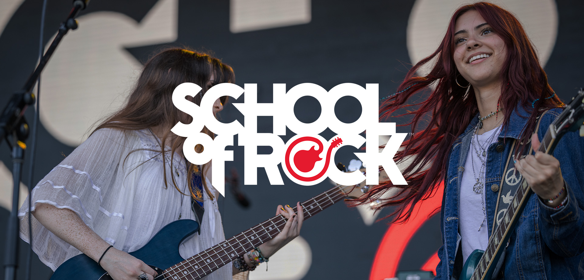 Two young women playing electric guitars on stage, one with long dark hair wearing a white blouse and the other smiling with red hair wearing a jean jacket, with a large 'School of Rock' logo overlay.