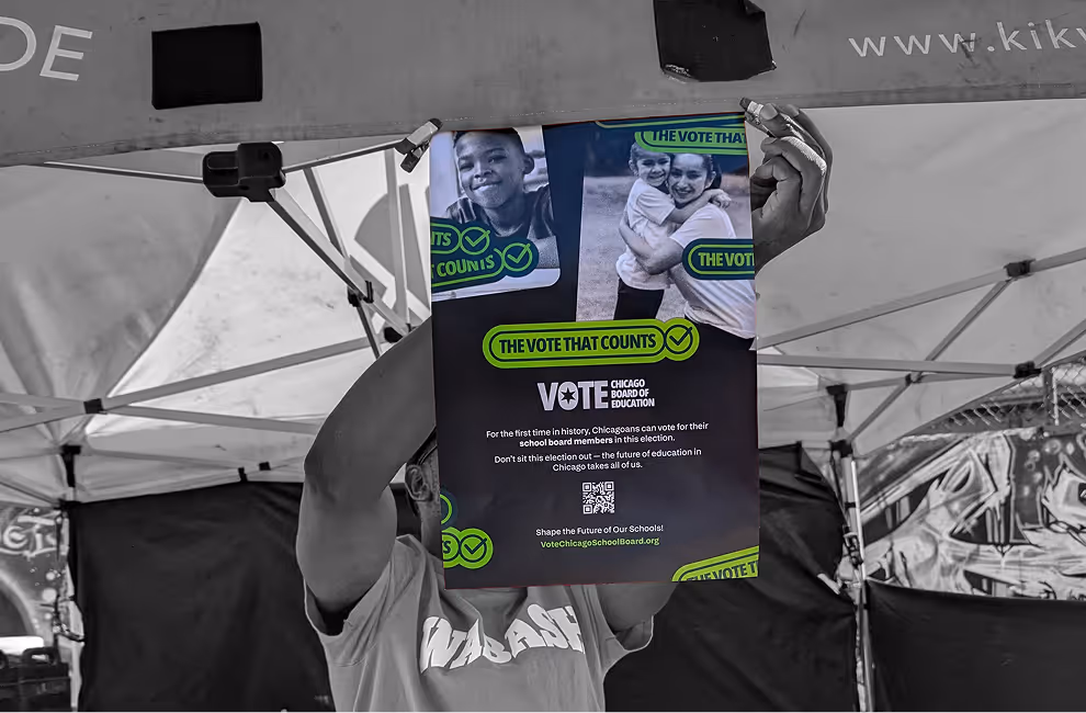 Person holding a colorful flyer under a tent with text encouraging Chicagoans to vote in the school board election.