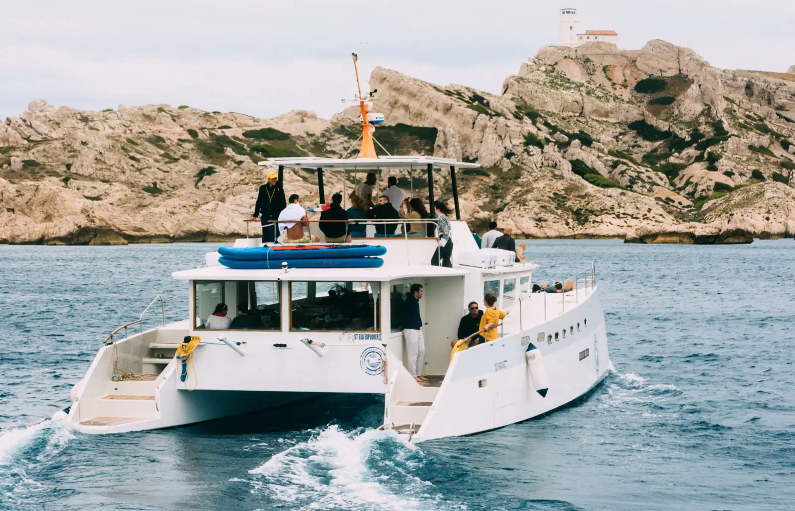 Bateau blanc avec plusieurs personnes à bord naviguant près des rochers avec un phare en arrière-plan.