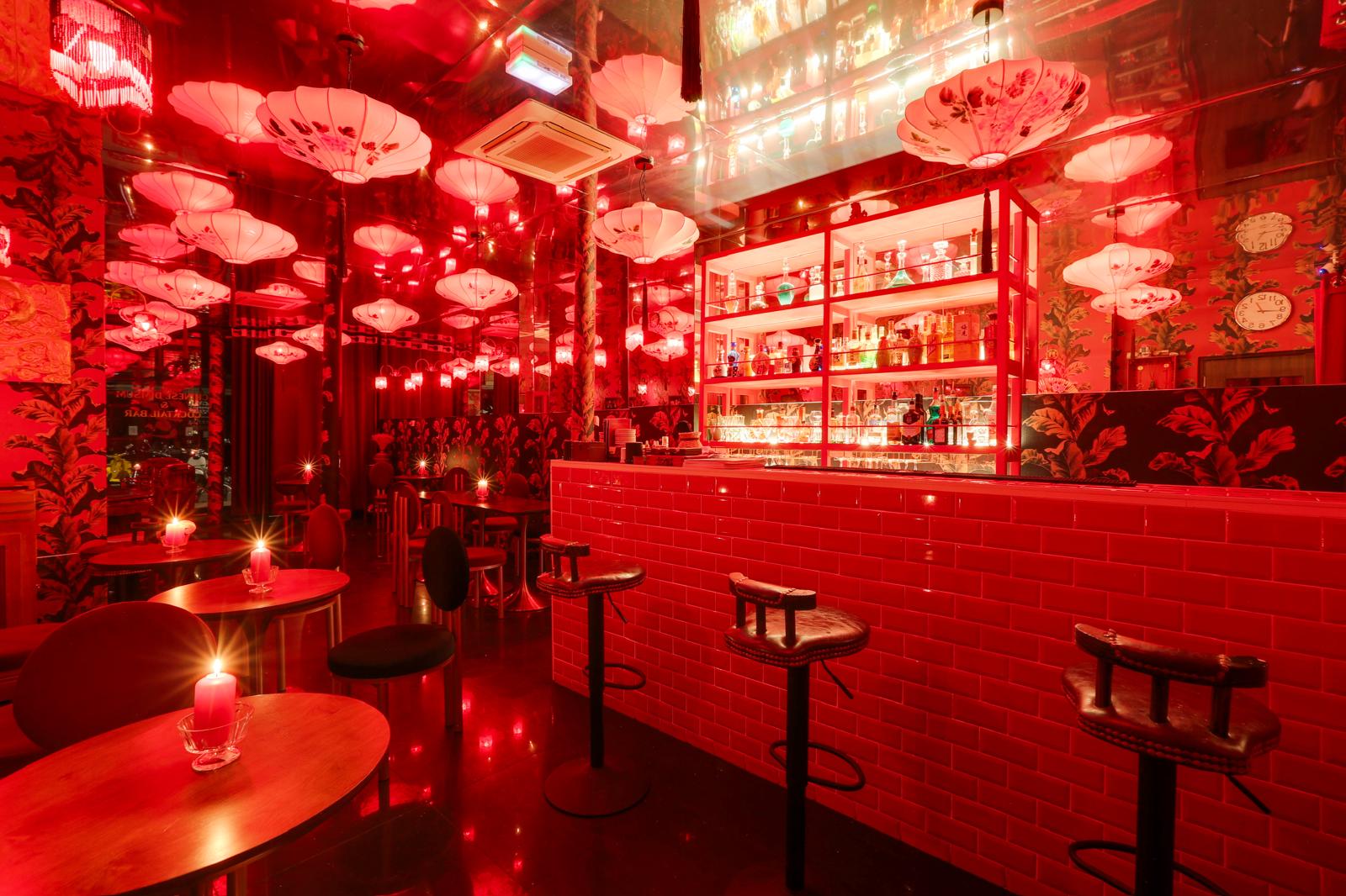 Red-lit bar interior with floral lanterns, red tiled bar, wooden tables, and black round stools.