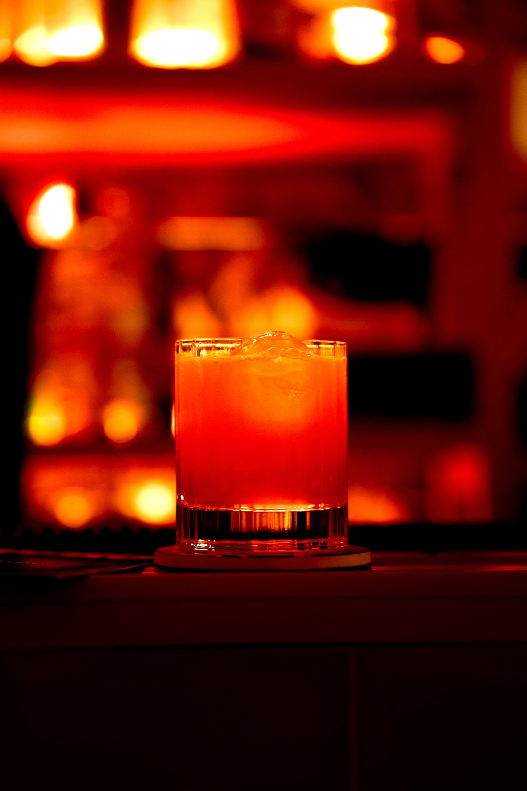 Close-up of an orange cocktail with ice in a clear glass on a bar with warm lighting.