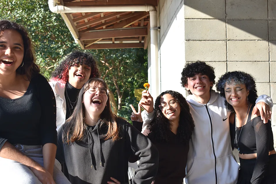 Group of six young people smiling and laughing outdoors against a wall and greenery, one person holding a small orange flower.
