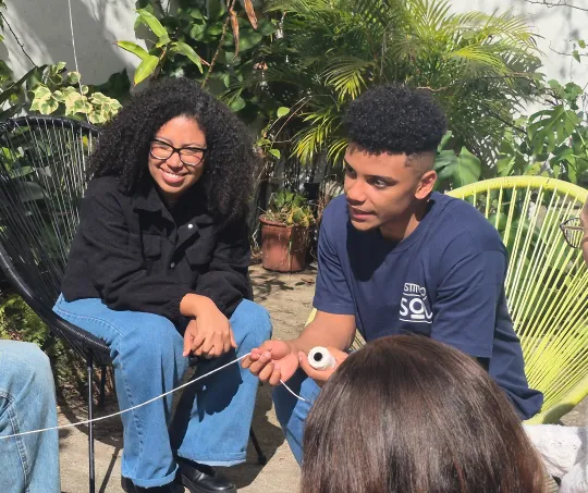 Two young people seated outdoors, one holding a spool of white string, surrounded by green plants and modern chairs.